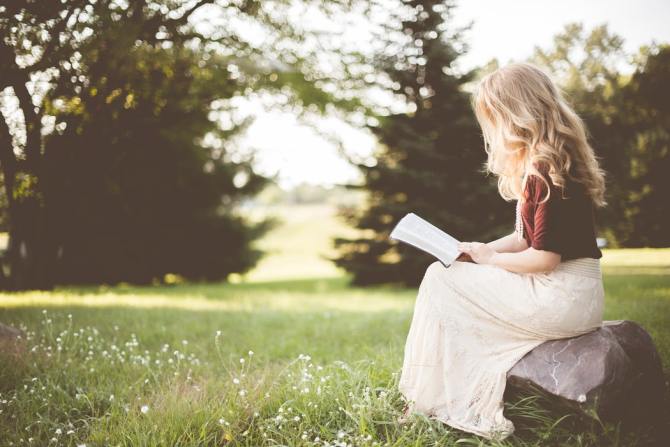 woman-with-blonde-hair-wearing-red-top-and-long-white-skirt-sitting-on-rock-in-meadow-while-reading-book