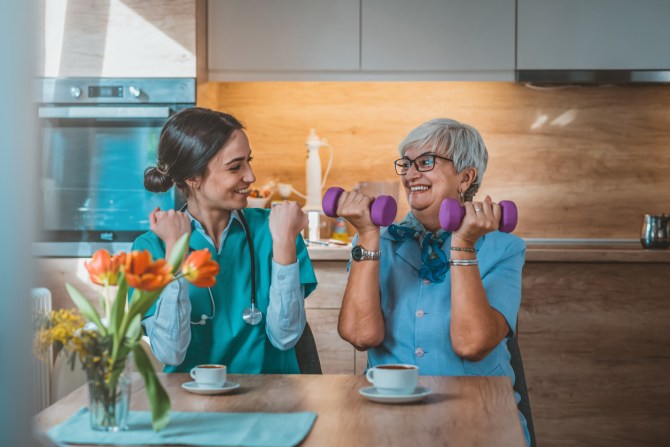 senior-woman-doing-dumbbell-curls-with-physical-therapist-at-kitchen-table-with-teacups-and-vase-of-orange-flowers