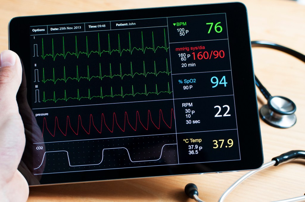 Doctor holding a digital tablet with a live recorded ecg on it. On the desk in the background is a stethoscope.