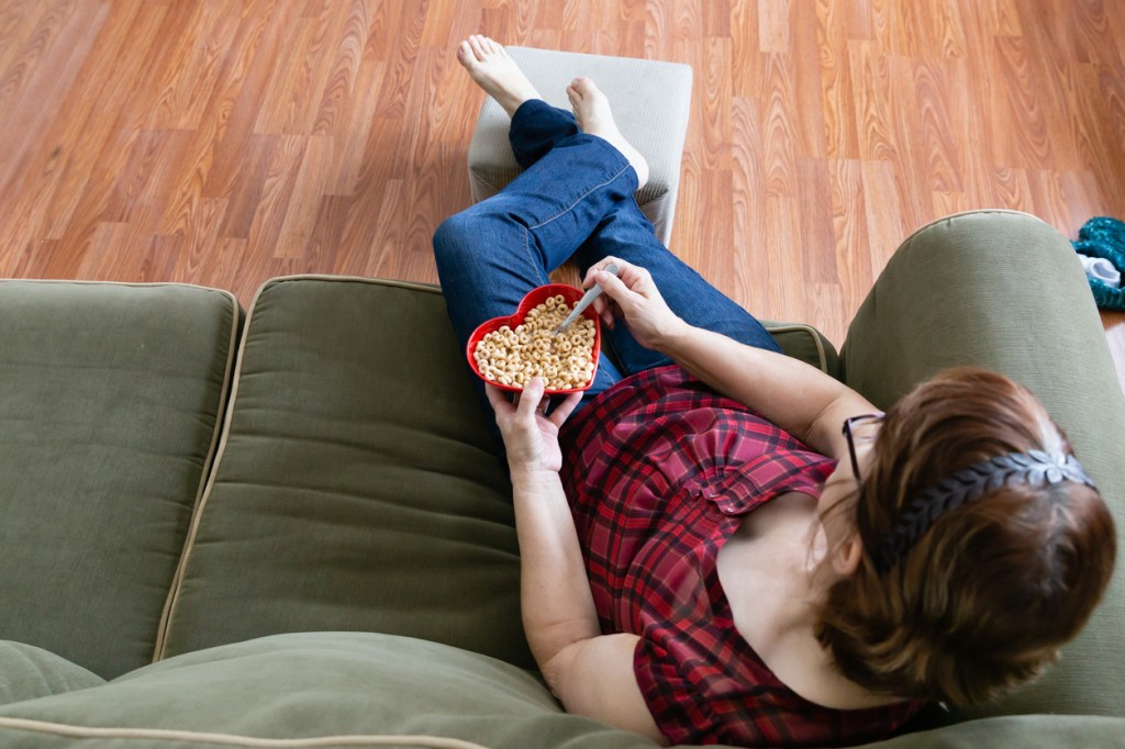 overhead-view-of-woman-sitting-on-sofa-with-feet-on-footrest-while-holding-heart-shaped-bowl-of-cheerios-with-spoon