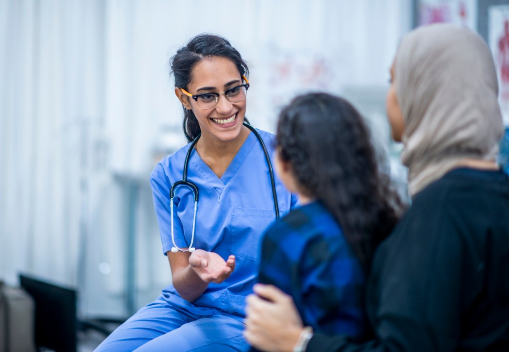 A mother and daughter speaking to a doctor who is wearing blue scrubs and a stethoscope during a doctor's appointment..