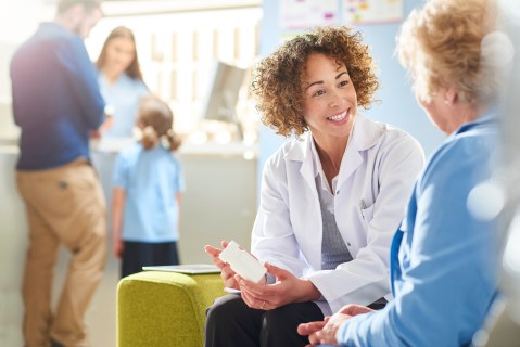 A female pharmacist sits with a senior female patient in the pharmacist consultation area and discusses her prescription and choice of medication. In the background a father and daughter stand at the dispensing counter and are served by a female pharmacy assistant .