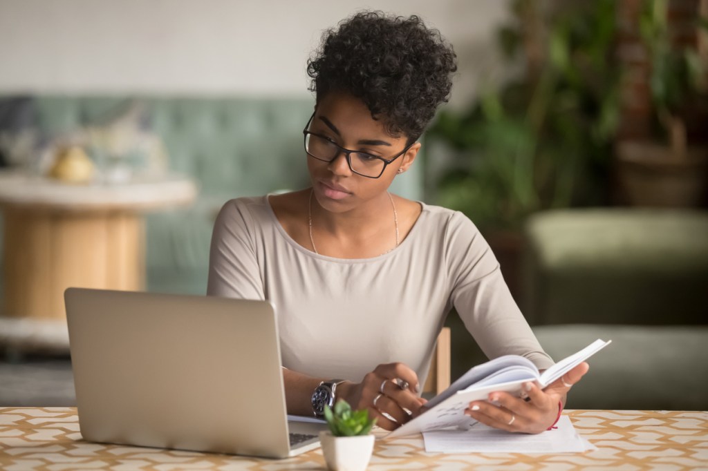 A young woman holding a book while sitting at a table and looking at a laptop while studying